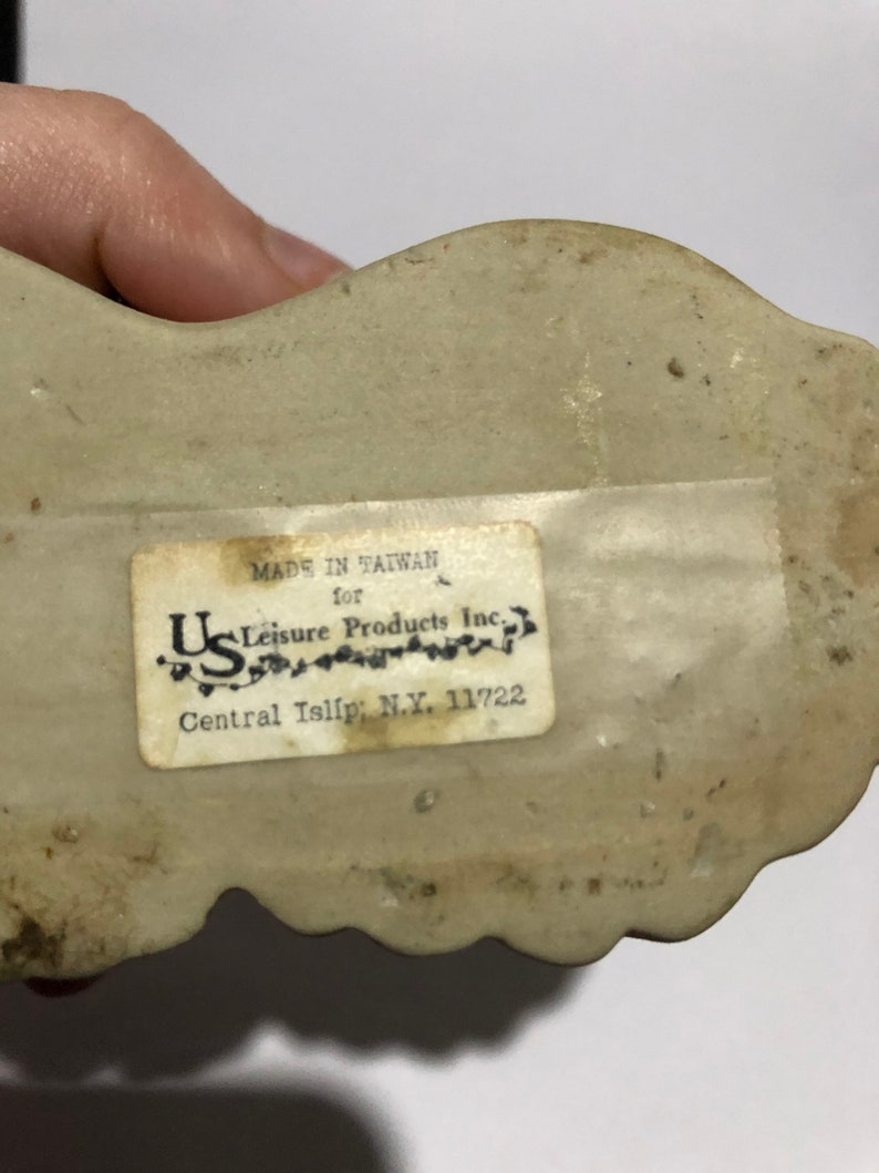 May include: A white ceramic figurine with a label that reads "Made in Taiwan for U.S. Leisure Products Inc. Central Islip, N.Y. 11722"