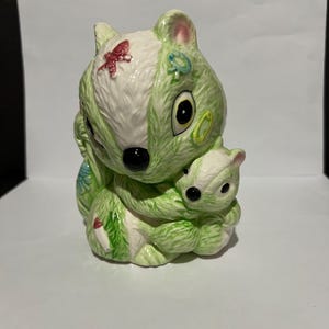 May include: A green and white ceramic figurine of a squirrel holding a smaller squirrel. The larger squirrel has a red butterfly, a blue flower, and green and yellow circles painted on its fur. The smaller squirrel has black eyes and a black nose.