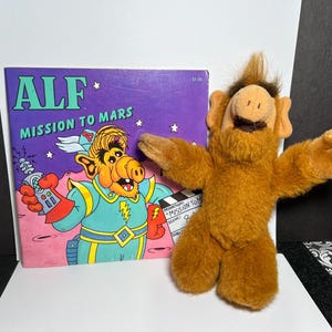 May include: A brown plush toy of the character ALF standing next to a square book titled ALF Mission to Mars. The book cover features a colorful illustration of the character in a space suit holding a ray gun against a purple starry background.
