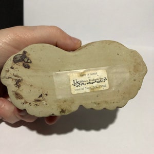 May include: A white ceramic figurine of a dog with a label on the bottom that reads "Made in Taiwan for U.S. Leisure Products Inc. Central Islip, N.Y. 11722"