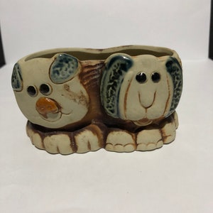 May include: A ceramic planter shaped like two dogs with brown, white, and blue glaze. The dogs are facing each other and have large eyes and a smiling mouth.