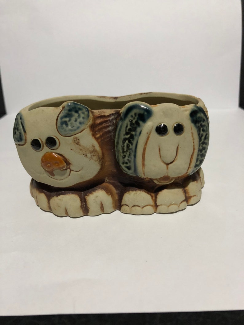 May include: A ceramic planter shaped like two dogs with brown, white, and blue glaze. The dogs are facing each other and have large eyes and a smiling mouth.
