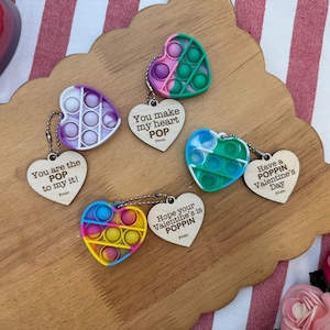 May include: Heart-shaped fidget toys in various colors, including purple, pink, blue, and yellow, with attached wooden tags. The tags have phrases like "You make my heart POP" and "Have a POPPIN Valentine's Day."