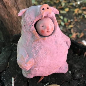 Può includere: Una statuetta scolpita a mano che presenta un volto rotondo dalla pelle pallida con guance rosate che spunta dal cappuccio di un costume da maiale rosa chiaro testurizzato. La figura ha un corpo robusto e arrotondato con finitura opaca.