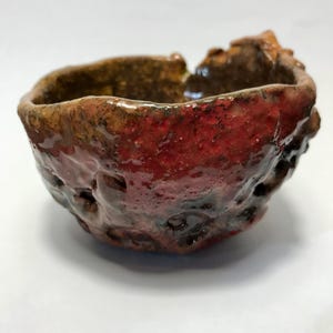 Può includere: Una piccola ciotola in ceramica formata a mano con una forma organica irregolare. L'esterno presenta una superficie ruvida e craterizzata, rifinita con uno smalto rosso lucido profondo che sfuma in toni scuri vicino alla base e al bordo.