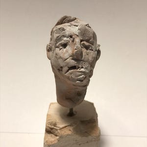 Puede incluir: Busto escultórico de una cabeza humana, elaborado con un material claro. El rostro presenta rasgos ásperos y texturizados, incluyendo nariz, boca y ojos. La cabeza está montada sobre una pequeña base rectangular de piedra, creando una exhibición artística única.