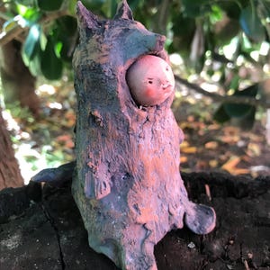 Può includere: Una piccola statuetta in argilla scolpita a mano che presenta un volto pallido e rotondo con guance rosate che spunta da un cappuccio testurizzato a forma di lupo, nei toni del grigio scuro e marrone terra, con orecchie a punta e superficie ruvida.