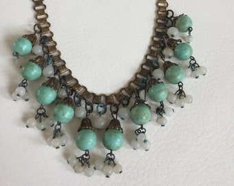 Peut inclure: Un collier en chaîne dorée avec des perles turquoise et blanches. Le collier a un design en cascade avec les perles turquoise qui pendent de la chaîne.