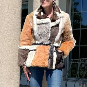 May include: A woman wearing a brown, white and black fur jacket with a brown leather belt. She is wearing blue jeans with a small hole in one knee and brown heeled shoes.