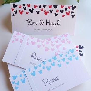 May include: A stack of rectangular white cards featuring colorful mouse-ear silhouettes at the top. The cards display custom text including Ben & Hollie Happy Honeymoon, Aurora, and Rome Disneyland Awaits, printed in black and gray fonts.
