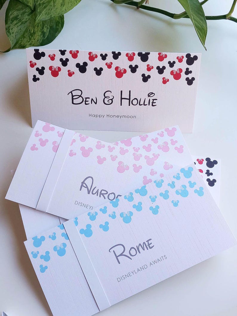 May include: A stack of rectangular white cards featuring colorful mouse-ear silhouettes at the top. The cards display custom text including Ben & Hollie Happy Honeymoon, Aurora, and Rome Disneyland Awaits, printed in black and gray fonts.