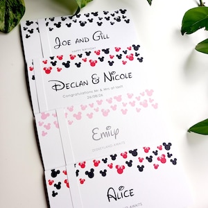 May include: Four white rectangular cards featuring black and red mouse-ear silhouettes. The cards display the names Joe and Gill, Declan and Nicole with the date 26/08/26, Emily, and Alice, along with phrases like Happy Birthday and Disneyland Awaits.