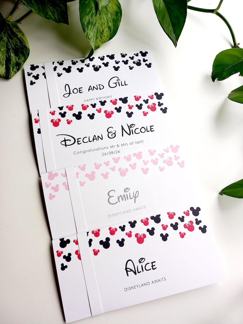 May include: Four white rectangular cards featuring black and red mouse-ear silhouettes. The cards display the names Joe and Gill, Declan and Nicole with the date 26/08/26, Emily, and Alice, along with phrases like Happy Birthday and Disneyland Awaits.