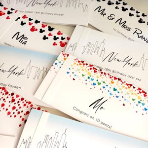 May include: A collection of rectangular white greeting cards featuring various printed designs including black New York city skylines, red and black mouse ear silhouettes, and colorful rainbow hearts, with personalized text such as names and dates.