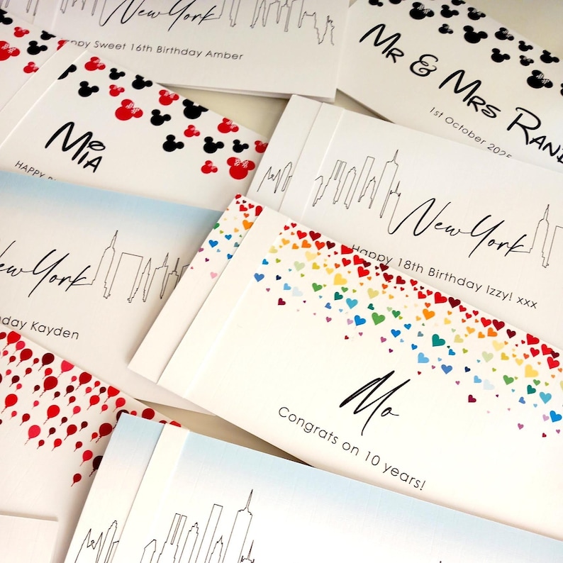 May include: A collection of rectangular white greeting cards featuring various printed designs including black New York city skylines, red and black mouse ear silhouettes, and colorful rainbow hearts, with personalized text such as names and dates.