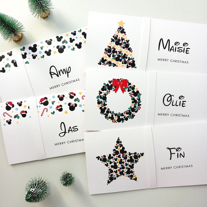 May include: A collection of white rectangular Christmas cards featuring personalised names including Amy, Jas, Maisie, Ollie, and Fin. The designs incorporate black mouse-ear silhouettes, red bows, candy canes, and festive shapes arranged into trees, wreaths, and stars.