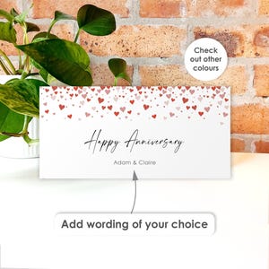 May include: A rectangular white card featuring a pattern of small red and pink hearts along the top edge. The text "Happy Anniversary" is printed in a black script font above the names "Adam & Claire" in a simple sans-serif typeface.