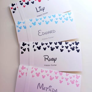 May include: Four white rectangular envelopes featuring a top border of scattered mouse-ear silhouettes. The envelopes are personalized with the names Lily, Edward, Ruby, and Matilda, each followed by the printed text Happy Easter.