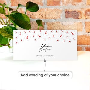May include: A white gift card with the name "Katie" in cursive and the text "DRIVING LESSON FUNDS". The card is decorated with red "L" symbols and the text "Add wording of your choice" at the bottom. A green plant and a brick wall are in the background.
