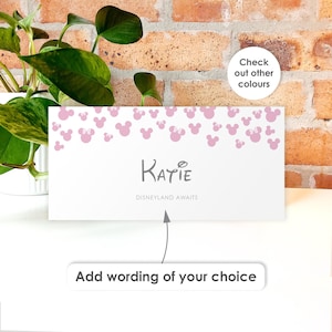 May include: A rectangular white card featuring a pattern of light pink Mickey Mouse silhouettes along the top edge. The center displays the name Katie in dark gray text, with the phrase Disneyland Awaits below it and an arrow pointing to the text.