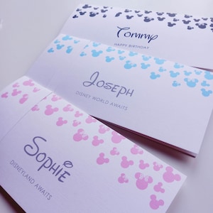 May include: Three rectangular white paper cards featuring mouse-ear silhouettes in dark blue, light blue, and pink. The cards display the names Tommy, Joseph, and Sophie, with the printed text HAPPY BIRTHDAY, DISNEY WORLD AWAITS, and DISNEYLAND AWAITS.