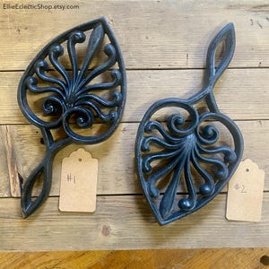 May include: Two cast iron trivets with a leaf-like design and a handle. The trivets are black and have an ornate, openwork pattern. Each trivet has a small tag attached with a number. The background is a wooden surface.