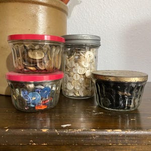 May include: Four glass jars with red, silver, and gold lids filled with various buttons. The jars are on a wooden surface.
