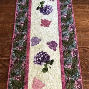May include: A quilted table runner with a light green center panel featuring embroidered purple hydrangeas and pink butterflies. The edges have a leaf pattern and pink trim, creating a decorative accent for a dining table or similar surface.