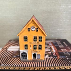 Puede incluir: Un edificio amarillo en miniatura con un tejado marrón, un reloj y varias ventanas. El edificio tiene las palabras "ENHJØRNINGEN" y "SJØBODEN" en la parte delantera. El edificio está sobre una superficie rayada.