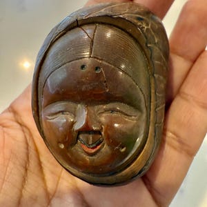May include: A carved wooden sculpture of a smiling face with closed eyes and a small open mouth. The sculpture is brown with a smooth, polished finish and appears to be an antique or vintage item. The face is framed by a head covering.
