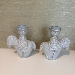 May include: Two matching ceramic candle holders. Each is shaped like a horse with a rider wearing a hat. The candle holders are light grey and have a glossy finish. The background is a textured, light beige wall.