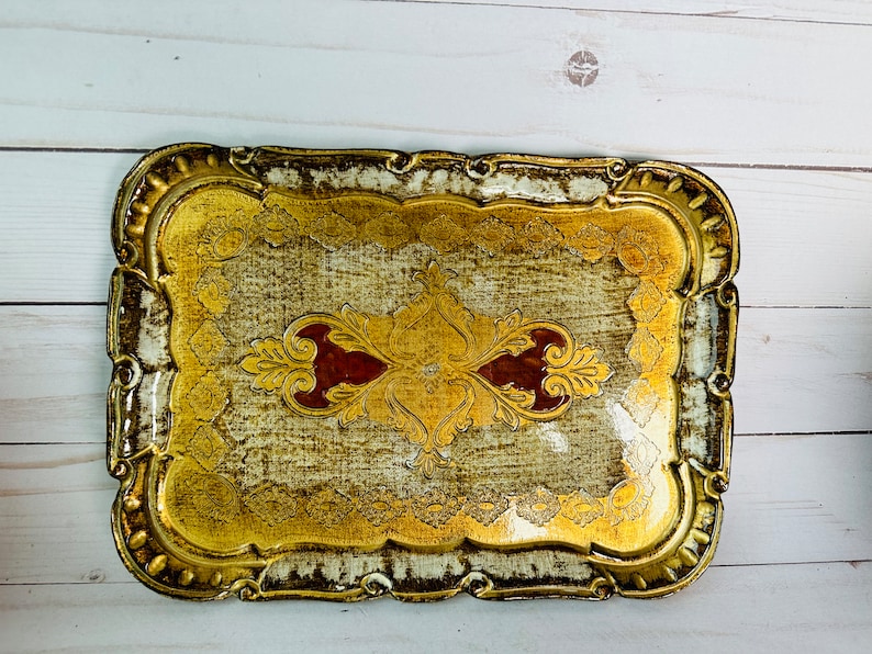 May include: A gold and white decorative tray with an ornate design. The tray has a scalloped edge and a raised center with a floral pattern.