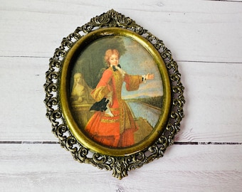 Puede incluir: Un marco de imagen ovalado de latón con un diseño ornamentado. El marco contiene una impresión vintage de una persona con un vestido rojo y dorado de pie en un paisaje.