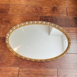 May include: Oval vanity mirror with a gold filigree frame. The mirror reflects a room with white walls and a light fixture. The frame has a decorative, repeating leaf pattern. The mirror is set on a wooden surface.