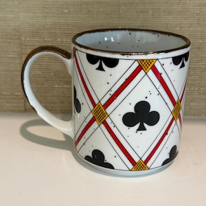 May include: White ceramic mug with a playing card club suit design. The mug features a repeating pattern of black clubs, red diagonal lines, and yellow diamond shapes. The rim and handle are brown.