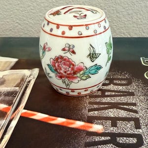 Puede incluir: Un pequeño tarro de cerámica blanca con tapa, decorado con motivos florales y mariposas pintados a mano en rojo, rosa, verde y azul. El tarro tiene forma cilíndrica y está adornado con lunares rojos.