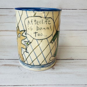 Puede incluir: Una taza de cerámica blanca con un borde azul. La taza tiene una ilustración dibujada a mano de dos figuras, una sosteniendo una copa de martini. El texto en la taza dice "Afterlife is banal, too..."