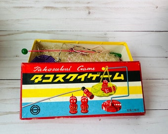 May include: Vintage Japanese Takosukui Game box with red, yellow, and blue stripes. The box contains game pieces, including small red figures and a balancing scale. The text "Takosukui Game" is printed on the box.