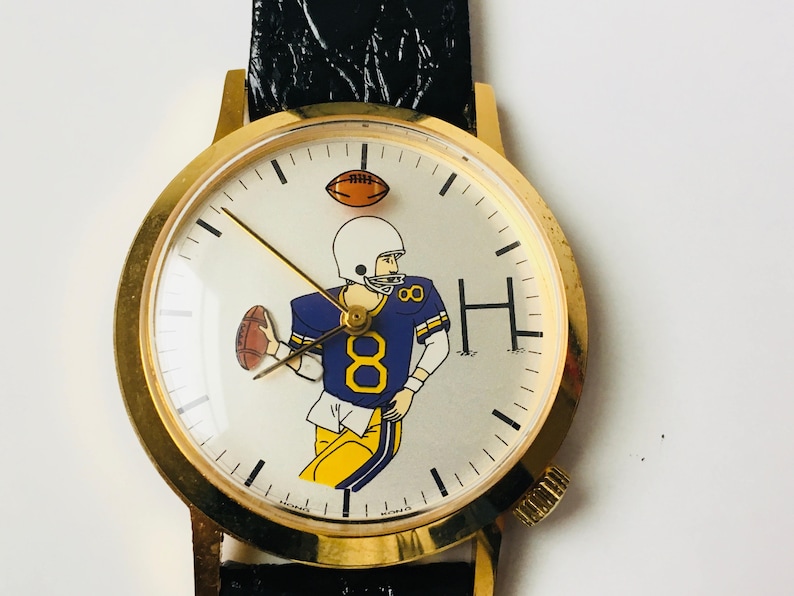 May include: Gold-tone wristwatch with a black leather band. The watch face features a cartoon illustration of a football player in a blue and yellow uniform, holding a football. The watch has a gold bezel and a white dial.