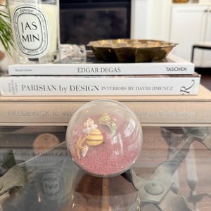May include: A glass globe filled with pink sand and seashells. The globe is sitting on a glass coffee table with a metal base.
