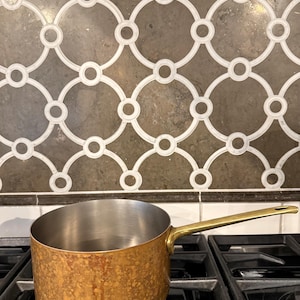 May include: A copper saucepan with a brass handle sits on a gas stove. The interior of the pan is stainless steel. The handle is angled and attached to the side of the pan. The background features a decorative tile backsplash.