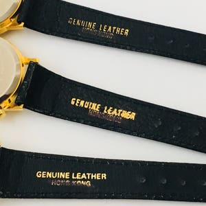May include: Close-up of three gold-toned watches with black leather straps. The watch backs are visible, and the straps are stamped with "GENUINE LEATHER HONG KONG" in gold lettering. The watches appear to be vintage.
