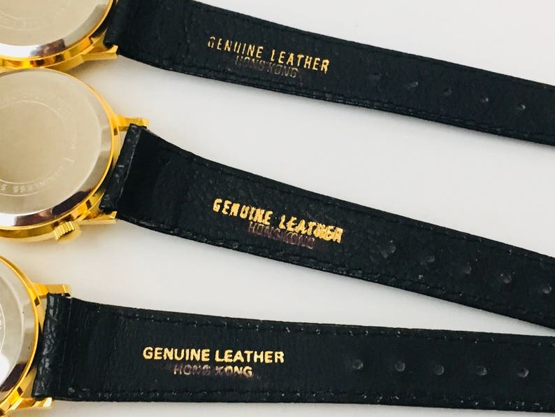 May include: Close-up of three gold-toned watches with black leather straps. The watch backs are visible, and the straps are stamped with "GENUINE LEATHER HONG KONG" in gold lettering. The watches appear to be vintage.
