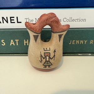 Puede incluir: Un pequeño jarrón de cerámica de color beige con una forma única y un asa de color marrón rojizo. El jarrón presenta un diseño geométrico negro y una figura con corona. El jarrón mide unos 7,6 cm de alto.