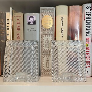May include: Two clear glass picture frames with a wavy, textured design sit on a white shelf in front of a collection of books. The books include titles such as "Crime and Punishment" and "David Copperfield".