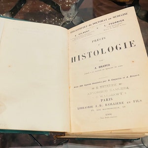 May include: A vintage book titled "Precis d'Histologie" by A. Branca. The book is bound in green leather and has a gold-embossed title on the cover. The book is open to the title page, which includes the author's name, the publisher's name, and the year of publication.
