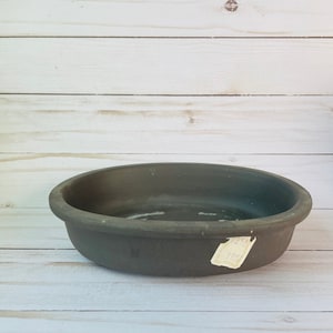 Peut inclure: Un bol en céramique ovale gris foncé avec un bord arrondi. Le bol est posé sur une surface en bois de couleur claire. Une petite étiquette rectangulaire est fixée sur le côté du bol.
