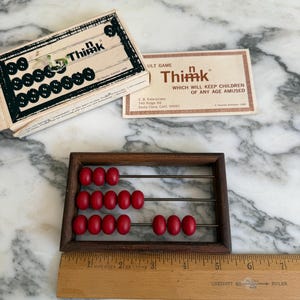 K&ouml;nnte beinhalten: Ein Vintage-Abakus aus Holz mit roten Perlen und einem Holzlineal. Die Spielbox und die Karte f&uuml;r "Think" sind ebenfalls sichtbar. Der Abakus hat einen dunkelbraunen Rahmen und Metallst&auml;be. Das Lineal ist in cm markiert.