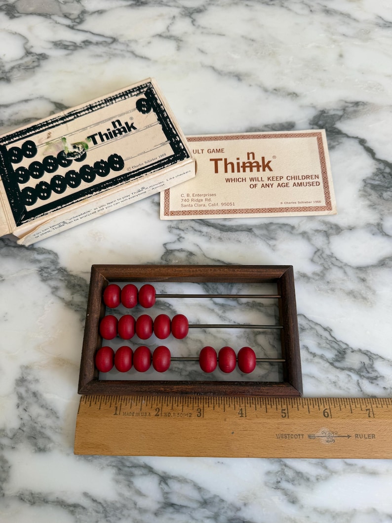 K&ouml;nnte beinhalten: Ein Vintage-Abakus aus Holz mit roten Perlen und einem Holzlineal. Die Spielbox und die Karte f&uuml;r "Think" sind ebenfalls sichtbar. Der Abakus hat einen dunkelbraunen Rahmen und Metallst&auml;be. Das Lineal ist in cm markiert.