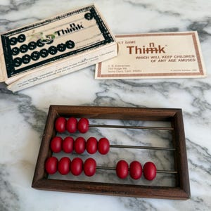 K&ouml;nnte beinhalten: Ein Vintage-Thimk-Spiel mit einem h&ouml;lzernen Abakus und einer Schachtel mit schwarzen Spielsteinen. Der Abakus hat einen dunklen Holzrahmen und rote Perlen. Die Spielbox und die Karte sind beige mit schwarzem und rotem Text.